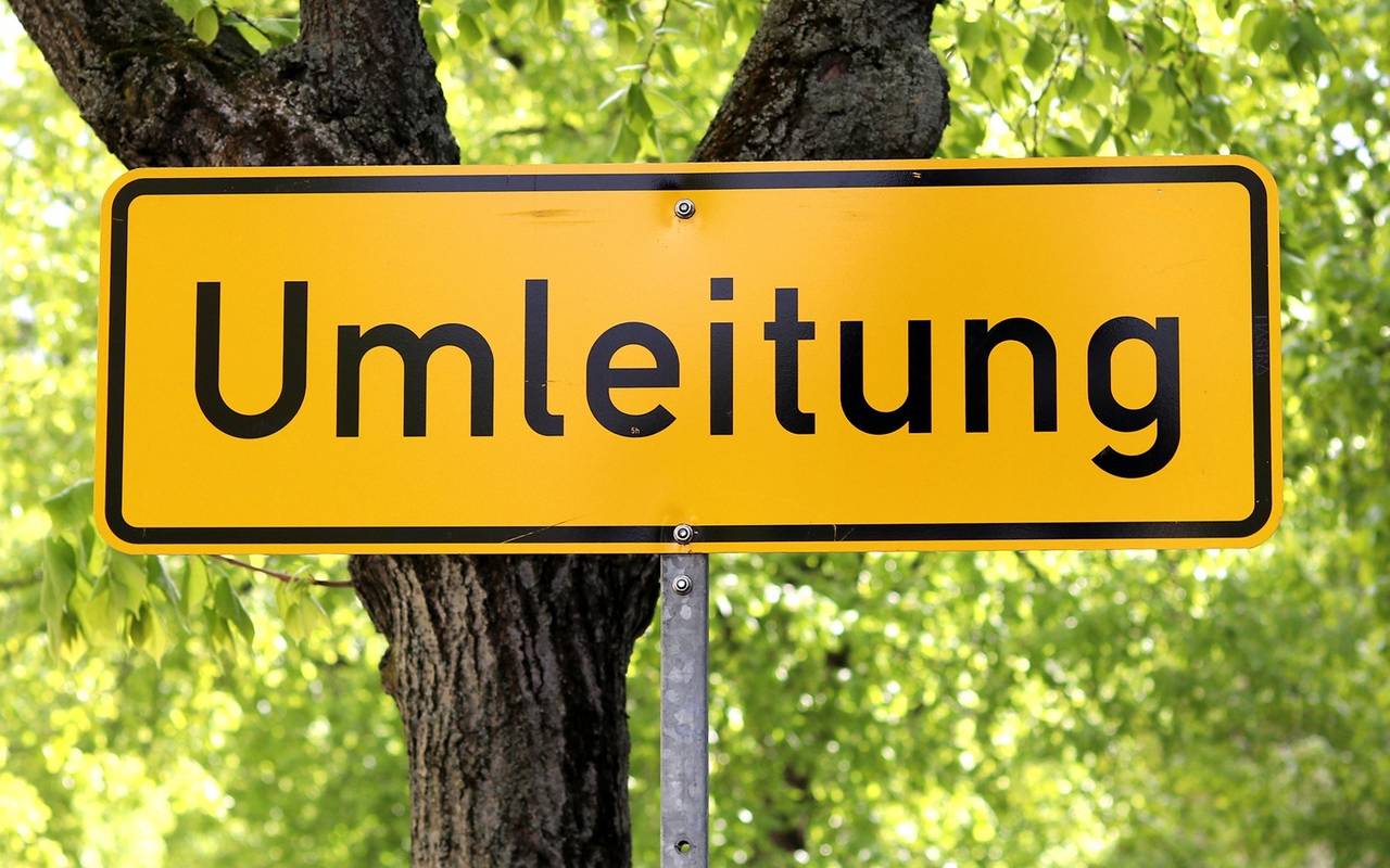 Ein Straßenschild aus Deutschland, das im Hintergrund gelb ist und die schwarze Aufschrift "Umleitung" trägt, steht vor einer Straßenkulisse mit einer begrünten Allee.