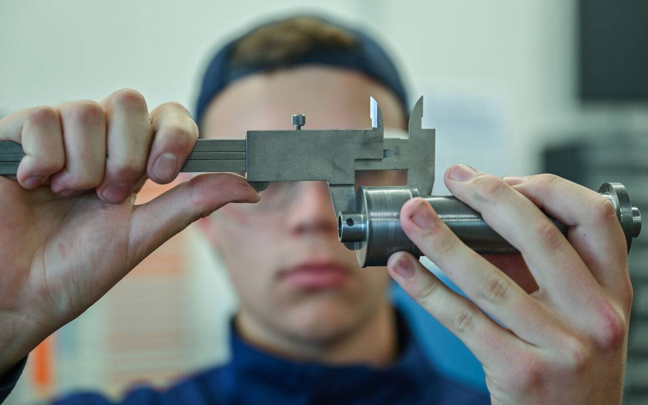 Ein Lehrling zum Mechatroniker prüft mit einem Messschieber ein Werkstück in einer Ausbildungsstätte