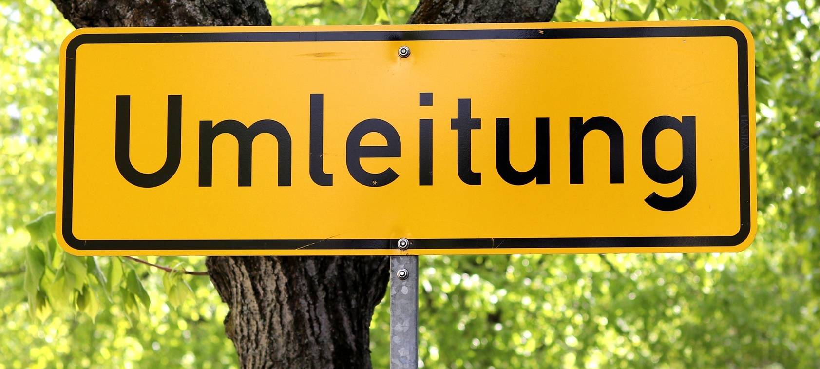 Ein Straßenschild aus Deutschland, das im Hintergrund gelb ist und die schwarze Aufschrift "Umleitung" trägt, steht vor einer Straßenkulisse mit einer begrünten Allee.