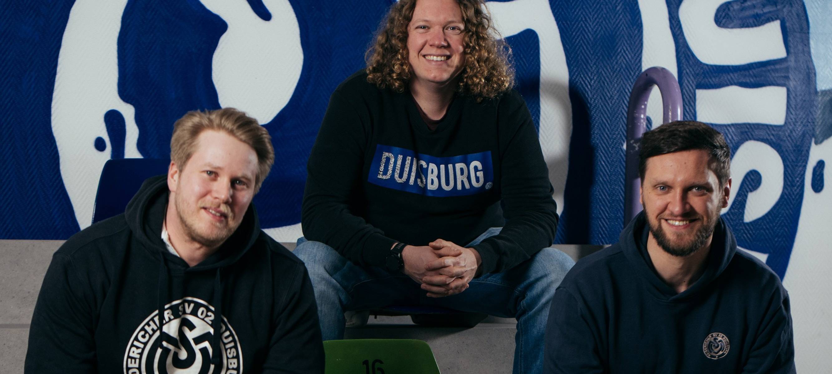 Die MSV-Fans Luca Joe Sankowski, Maximilian Winkel und Thomas Biehl-Wisniewski sitzen gemeinsam vor einem großen MSV-Logo im Vereinsheim der Zebras.