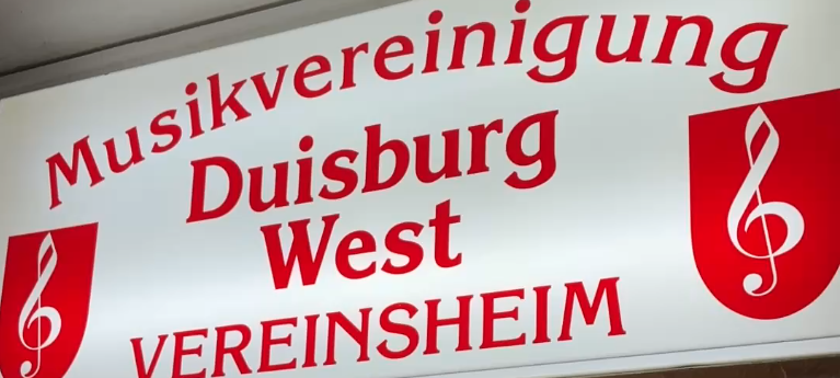 Ein beleuchtetes Schild zeigt in roter Schrift auf weißem Grund das Logo der Musikvereinigung Duisburg West